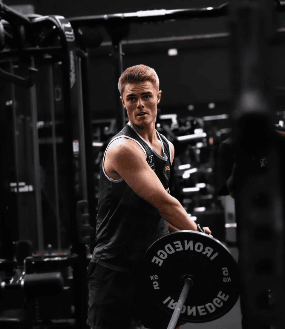 A blond man in a black tank top lifting a barbell in a gym setting, with workout machines in the background.